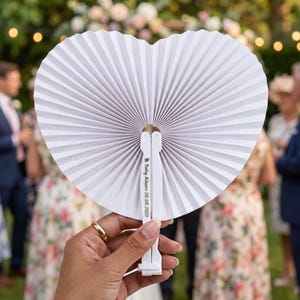Peut inclure: Éventail en papier blanc en forme de cœur avec un design plissé. Le ventilateur est maintenu ouvert, révélant une poignée avec le texte "Baby Adams 26.08.2021". L'arrière-plan montre une réunion en plein air floue avec des personnes.