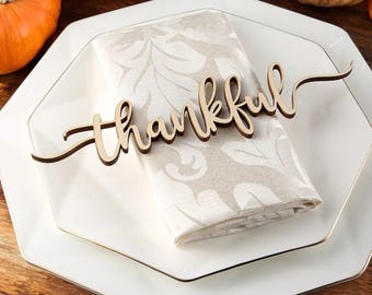 Könnte beinhalten: Ein weißer achteckiger Teller mit goldfarbenem Rand, eine gefaltete weiße Serviette mit beigefarbenem Blumenmuster und ein Holzschild mit der Aufschrift "thankful".