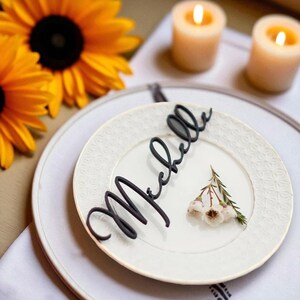 May include: A white plate with a floral pattern features a sprig of white flowers and a black cursive nameplate reading "Michelle." Two lit candles and sunflowers are in the background, creating a warm atmosphere.