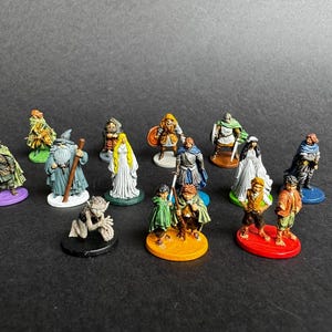 Può includere: Una collezione di quattordici miniature fantasy dipinte a mano, posizionate su basi circolari individuali di vari colori. Le figure includono guerrieri in armatura, maghi in tunica, viaggiatori con mantello e piccole creature con abbigliamento dettagliato.