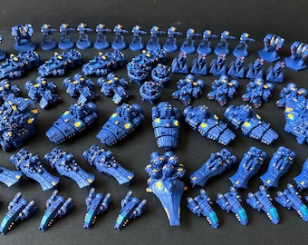 May include: A collection of blue and gold painted miniature spaceships and fighters. The miniatures are arranged in rows on a dark blue surface.
