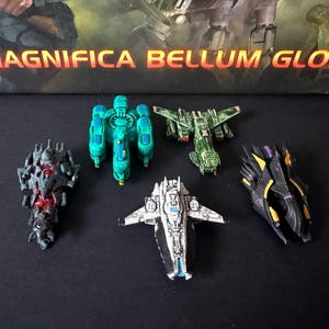 Puede incluir: Una colección de modelos de naves espaciales en miniatura en varios colores, incluyendo verde, negro y blanco. Los modelos están dispuestos sobre una superficie oscura, con el texto "MAGNIFICA BELLUM GLORIA" visible en el fondo.