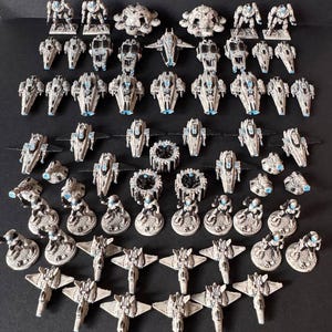 Puede incluir: Una colección de modelos de naves espaciales en miniatura de varios diseños. Los modelos son principalmente blanquecinos con detalles azules y negros. El surtido incluye cazas, robots y otros vehículos futuristas.