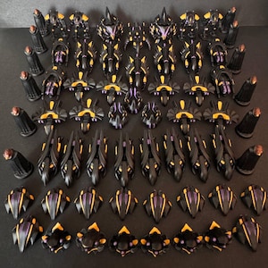 Könnte beinhalten: Eine Sammlung schwarzer und gelber Miniaturfiguren mit lila Akzenten. Die Figuren sind in Reihen und Spalten angeordnet und zeigen verschiedene Formen und Designs. Einige Figuren haben eine hohe, turmartige Struktur, während andere kompakter sind.