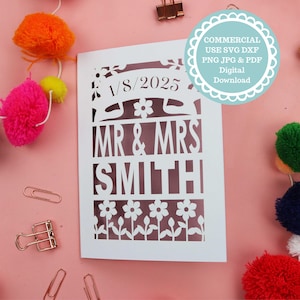 May include: White card with a cut-out design featuring the text "1/8/2025", "MR & MRS", and "SMITH". The design also includes small flowers and leaves. The card is on a pink background.