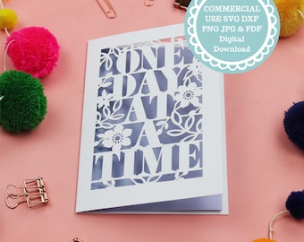 May include: White card with a cut-out design of the words "One Day At A Time" with floral accents. The card is on a pink background with colourful pom-poms.