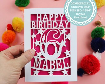 Op de afbeelding: Een witte, lasergesneden verjaardagskaart met de woorden "Happy Birthday", het nummer "6" en de naam "Mabel". De kaart wordt tegen een roze achtergrond gehouden, met kleurrijke pompons op de achtergrond.