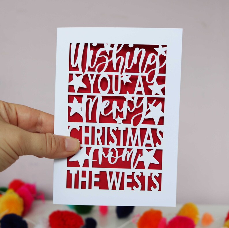 K&ouml;nnte beinhalten: Wei&szlig;e Karte mit rotem Hintergrund und einem ausgeschnittenen Design, das "Wishing you a Merry Christmas from the Wests" lautet.