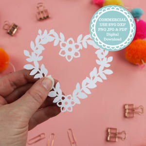 Puede incluir: Corte de papel blanco de una corona en forma de corazón con hojas y flores. La corona se sostiene en una mano sobre un fondo rosa. El texto "COMMERCIAL USE SVG DXF PNG JPG & PDF Digital Download" está en un círculo en el lado derecho de la imagen.