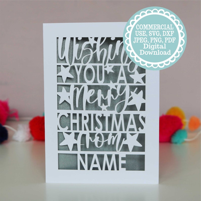 K&ouml;nnte beinhalten: Wei&szlig;e Karte mit einem ausgeschnittenen Design, auf dem "Wishing you a Merry Christmas from NAME" steht. Die Karte hat einen wei&szlig;en Hintergrund und der Text ist schwarz. Die Karte ist von bunten Pompons umgeben.