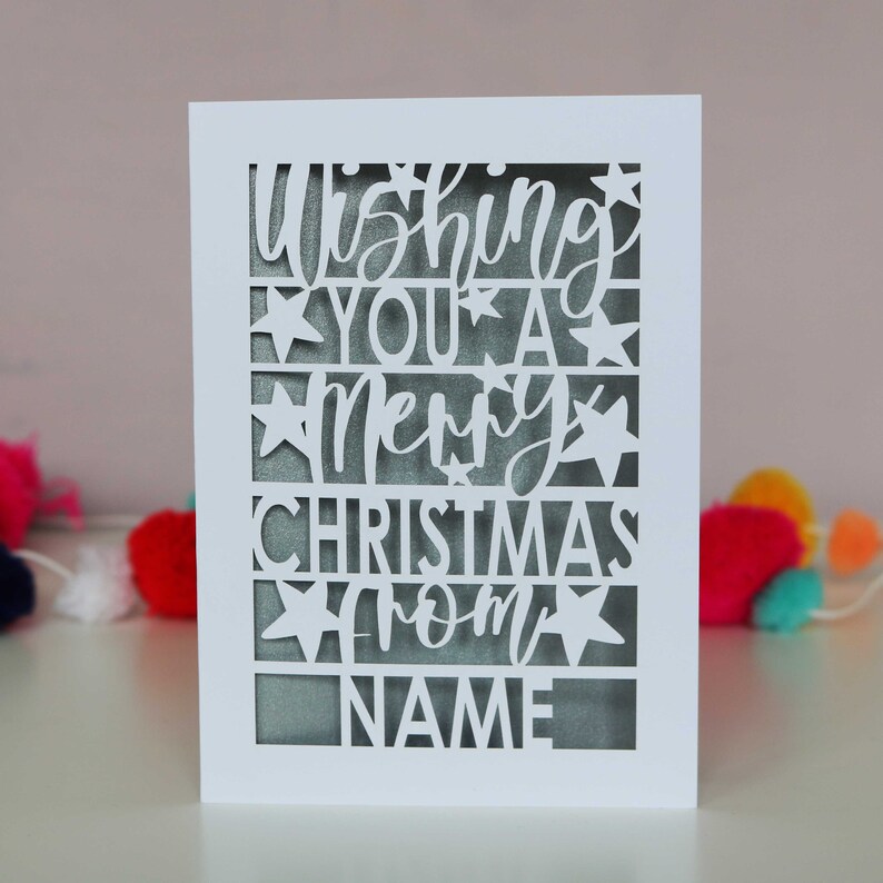 K&ouml;nnte beinhalten: Wei&szlig;e Karte mit einem ausgeschnittenen Design, auf dem "Wishing you a Merry Christmas from NAME" steht.