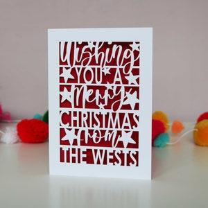 K&ouml;nnte beinhalten: Wei&szlig;e Karte mit rotem Hintergrund und einem ausgeschnittenen Design, das "Wishing you a Merry Christmas from the Wests" lautet.