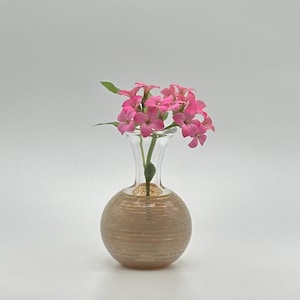 May include: A small, clear glass vase with a round, textured, tan base and a slender neck, holding a cluster of vibrant pink flowers with green leaves. The vase is a decorative item, perfect for displaying small floral arrangements.