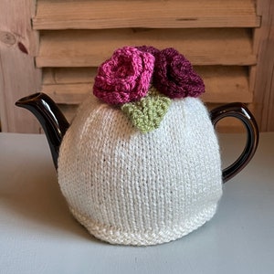 May include: White knitted teapot cosy with a pink and burgundy crocheted flower design.