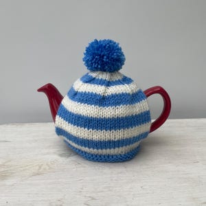 May include: A blue and white striped knitted teapot cosy with a blue pom pom on top. The cosy is on a red teapot.