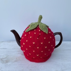 May include: A red knitted teapot cosy shaped like a strawberry with green leaves. The cosy is on a brown teapot.