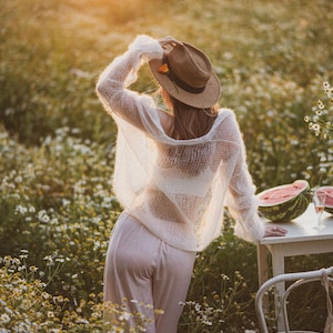 Könnte beinhalten: Eine Person in einem Feld mit Wildblumen, die einen weißen, durchsichtigen Pullover, eine hellrosa Hose und einen braunen Hut trägt. Ein Tisch mit Wassermelone und einem Glas Wein ist in der Nähe. Die Szene ist von warmem Sonnenlicht durchflutet.