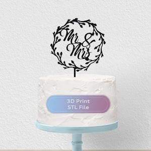 May include: Black 3D printed cake topper with a wreath and the words "Mr & Mrs" in a cursive font. The topper is on a white cake on a light blue cake stand. 3D Print STL File.