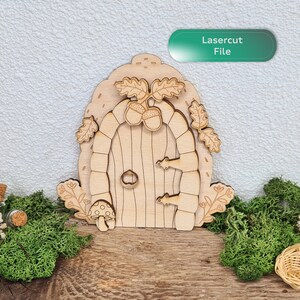 May include: A laser cut wooden fairy door with a rounded archway, a detailed door with a knob and hinges, and leaves and acorns on the top. The door is on a wooden surface with moss on either side.