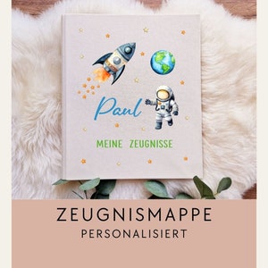 Könnte beinhalten: Ein Album aus beigefarbenem Leinen mit einer Aquarell-Illustration einer Rakete, eines Astronauten und der Erde. Das Album trägt den Namen "Paul" und den Text "Meine Zeugnisse" aufgedruckt. Das Album liegt auf einer weißen, flauschigen Oberfläche.