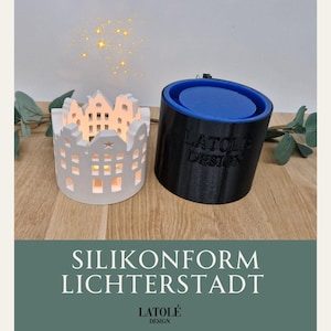 Könnte beinhalten: Zwei dekorative Kerzenhalter. Einer ist weiß, geformt wie eine Reihe von Häusern mit brennenden Kerzen im Inneren. Der andere ist ein schwarzer Zylinder mit blauem Innenraum. Der Text "LATOLÉ DESIGN" ist auf dem schwarzen Zylinder sichtbar. Der Text "SILIKONFORM LICHTERSTADT" ist ebenfalls sichtbar.