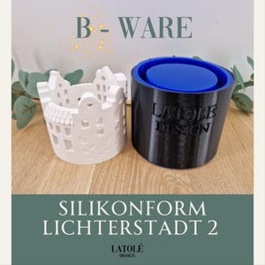 Peut inclure: Deux moules décoratifs. L'un est blanc, en forme de ville miniature avec des fenêtres découpées et des détails floraux. L'autre est noir avec un intérieur bleu. Le texte sur l'image indique "B-WARE", "SILIKONFORM LICHTERSTADT 2" et "LATOLÉ DESIGN".