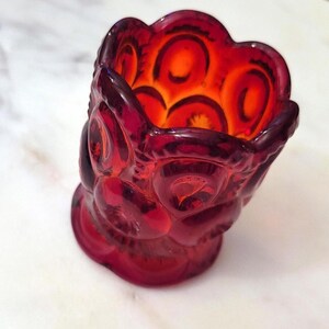 May include: A small, red glass dish with a scalloped rim and a starburst pattern. The dish is made of ruby red glass and has a clear, slightly orange hue.
