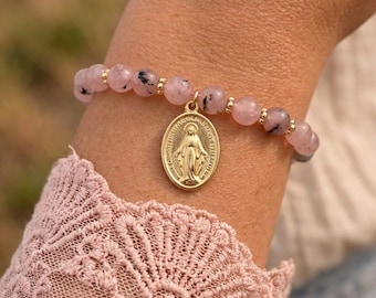 May include: A beaded bracelet featuring round, pale pink stones with dark inclusions, interspersed with small gold-toned metal beads. An oval gold-toned pendant depicting a figure with outstretched arms hangs from the center of the bracelet.