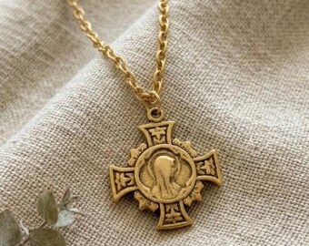 May include: A gold-colored pendant necklace featuring a cross design with an embossed image of a religious figure. The pendant hangs from a delicate chain. The necklace is displayed on a neutral-colored fabric.
