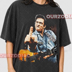 Puede incluir: Camiseta negra con una imagen desgastada de un hombre tocando la guitarra y mostrando el dedo medio. La imagen incluye el texto "OURZODIA" en blanco. El hombre lleva una camisa vaquera azul.