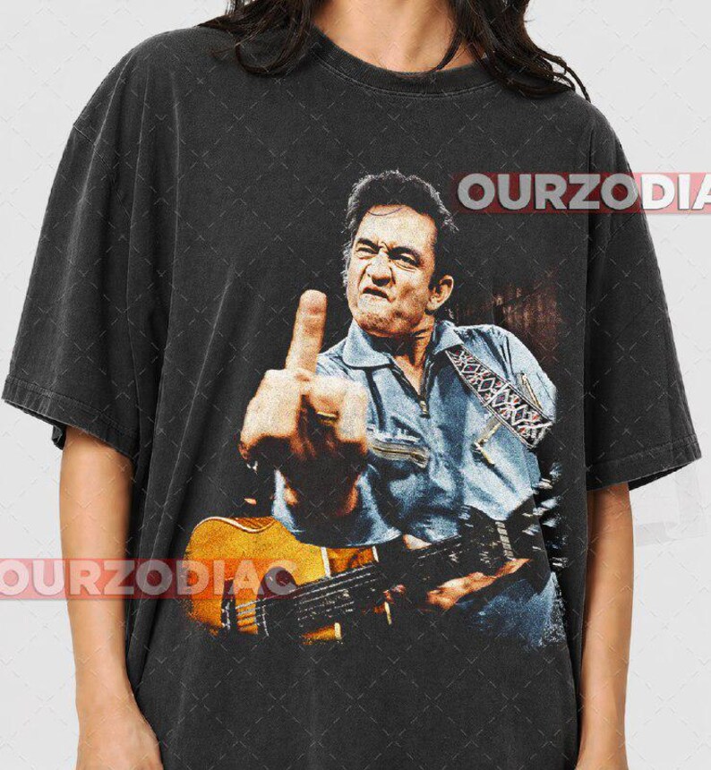 Puede incluir: Camiseta negra con una imagen desgastada de un hombre tocando la guitarra y mostrando el dedo medio. La imagen incluye el texto "OURZODIA" en blanco. El hombre lleva una camisa vaquera azul.