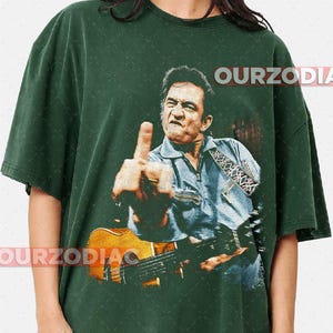 Puede incluir: Camiseta verde oscuro con una imagen vintage de un hombre con una guitarra, mostrando el dedo coraz&oacute;n. La imagen es de estilo desgastado. El texto "OURZODIA" est&aacute; impreso en la parte superior e inferior de la camiseta.