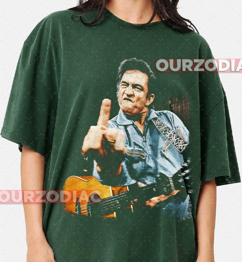 Puede incluir: Camiseta verde oscuro con una imagen vintage de un hombre con una guitarra, mostrando el dedo coraz&oacute;n. La imagen es de estilo desgastado. El texto "OURZODIA" est&aacute; impreso en la parte superior e inferior de la camiseta.
