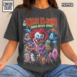 Op de afbeelding: Een antracietgrijs t-shirt met een grafische print van de film Killer Klowns from Outer Space. Het ontwerp bevat een centrale clownfiguur met een laserpistool, een bak popcorn, een vliegende schotel en de tekst Killer Klowns from Outer Space.