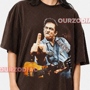 Puede incluir: Camiseta marr&oacute;n con una imagen desgastada de un hombre tocando la guitarra y mostrando el dedo medio. La imagen incluye el texto "OURZODIA". El hombre lleva una camisa vaquera azul.