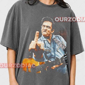 Puede incluir: Camiseta gris oscuro con una imagen desgastada de un hombre tocando la guitarra y mostrando el dedo medio. La imagen es en tonos azules, marrones y naranjas. El texto "OURZODIAC" est&aacute; impreso en la camiseta.