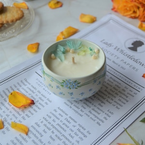 Peut inclure: Une bougie blanche avec des fleurs séchées sur le dessus, dans un récipient en céramique bleu et blanc. La bougie est posée sur une page d'un livre intitulé "Lady Whistledown's Society Papers".