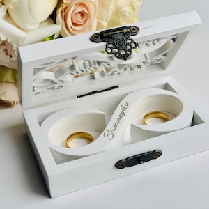 May include: A white wooden ring box with a hinged lid featuring an ornate dark metal latch. Inside, a white infinity-shaped insert holds two gold bands. The insert is engraved with Cyrillic text. The box is positioned near a bouquet of pale roses.