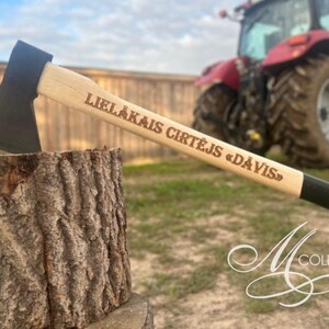 Puede incluir: Un hacha de mano con cabeza de metal negro y mango de madera clara que presenta el texto grabado LIELĀKAIS CIRTĒJS «DĀVIS». El hacha está incrustada en un tronco de textura rugosa. El mango tiene una empuñadura negra en el extremo.