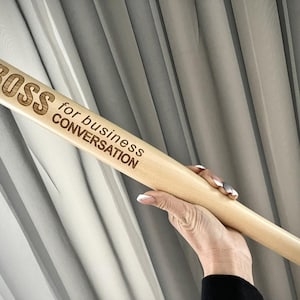 Könnte beinhalten: Ein Baseballschläger aus hellem Holz mit den Worten "BOSS for business CONVERSATION" auf dem Lauf eingraviert. Der Schläger wird von einer Person gehalten. Der Hintergrund ist ein hellgrauer Vorhang.