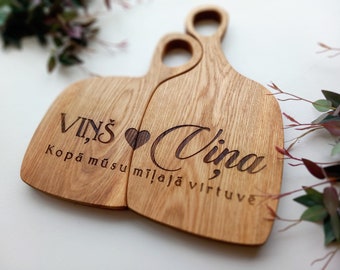 May include: Two wooden cutting boards with a handle, featuring engraved text and a heart symbol. The boards are light brown and have the words "Viņš" and "Vina" with additional text. A decorative kitchen item.