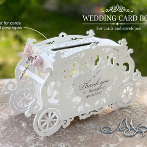 Könnte beinhalten: Eine weiße, lasergeschnittene Hochzeitskartenbox in Kutschenform mit filigranen Schnörkeln, vier Rädern und einem Schlitz oben. Eine herzförmige Plakette an der Seite trägt die Aufschrift Thank you for sharing our special day! Eine blassrosa Schleife.