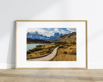 May include: A winding dirt road leads through a grassy valley towards a mountain range with snow-capped peaks and a blue lake in the background.