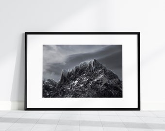 May include: A black and white photograph of a mountain range with a dramatic sky. The mountain peaks are jagged and snow-covered, and the clouds are dark and swirling.