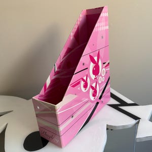 May include: Pink magazine file with a design featuring the Playboy bunny logo and geometric shapes. The file has a slanted design and the word "Playboy" is printed on the front.