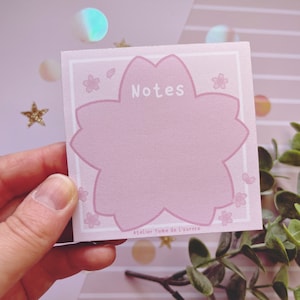 Könnte beinhalten: Ein rosa Haftnotiz mit einem Kirschblüten-Design. Die Notiz hat einen großen, blütenförmigen zentralen Bereich zum Schreiben, mit dem Wort "Notes" in Weiß. Kleine Kirschblüten-Illustrationen sind an den Rändern verstreut. Der Text "Atelier Yume de l'Aurore" befindet sich unten.