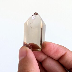 May include: A clear, faceted crystal point with a light brown tint. The crystal is held in a hand, highlighting its polished surfaces and natural variations. The crystal's tip is pointed, and the base has a darker, more opaque appearance.