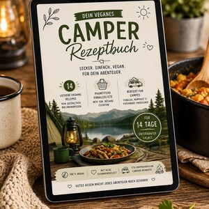 May include: A tablet displaying a vegan camper recipe book. The cover features the title "CAMPER Rezeptbuch" in green, with illustrations of camping scenes and food. The tablet is on a wooden surface, with a mug and lantern nearby.