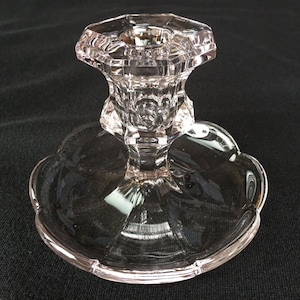 May include: A clear, faceted glass candlestick holder with a scalloped base. The candlestick has a hexagonal top and a detailed stem. The glass reflects light, creating a sparkling effect. The overall design is elegant and classic.