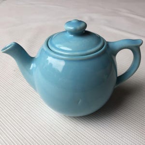 May include: A light blue ceramic teapot with a matching lid and handle. The teapot has a rounded body, a curved spout, and a small knob on the lid. The teapot is designed for brewing tea.